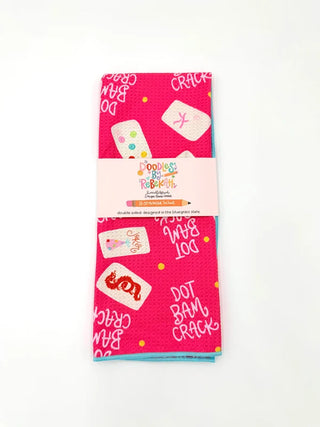 A folded pink cotton tea towel with a white paper band label in the center. The towel features a playful pattern of white bingo cards with colorful dots, hand-drawn style text like 'CRACK' and 'DOT BAML CRACK', and small yellow polka dots.