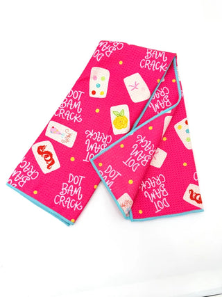 A folded bright pink fabric item, likely a scarf or napkin, featuring a playful print of white dominoes, a yellow pineapple, red cherries, and the words 'DOT BAM CRACK' in white script, with a light blue edge trim, displayed on a clean white background.