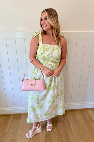 Woman in a floral dress with a pink handbag against a white paneled wall.