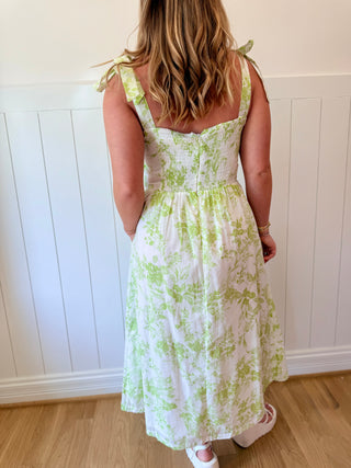 Woman wearing a white dress with green floral pattern against a white paneled wall.