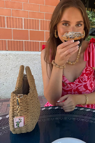 Woman in a red floral dress holding a pastry next to a straw bag with a tag.