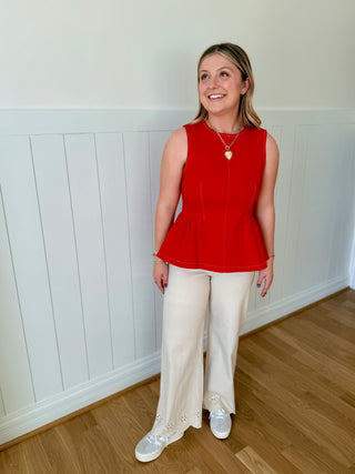 Woman wearing a red sleeveless top and white pants standing in a room with wooden flooring and white walls.