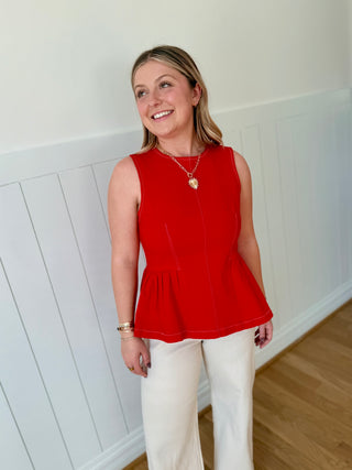 Woman wearing a red sleeveless top and white pants standing against a white paneled wall.
