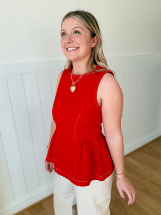 Woman wearing a red sleeveless top and white pants indoors.