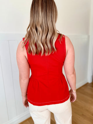 Person wearing a red sleeveless top and white pants in a room with wooden flooring.