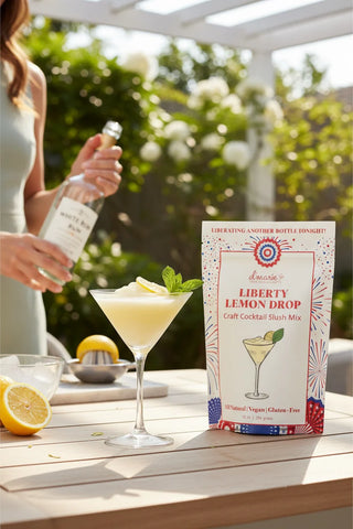 Liberty Lemon Drop cocktail mix box with a cocktail and lemon on a table outdoors.