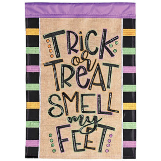 A rectangular decorative garden flag with a burlap-textured center panel reading “Trick or Treat Smell my Feet” in stylized lettering. The top and bottom borders are black with alternating pastel stripes in purple, pink, yellow, green, and white.