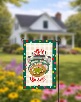 Decorative garden flag with race track design and 'Off to the Races' text, set against a blurred house and garden background.