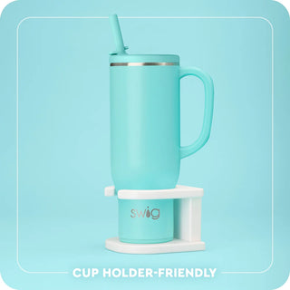 A mint green insulated travel mug with a handle, lid, and straw, displayed on a white cup holder stand. The mug features the 'swig' brand logo in gold on the holder.