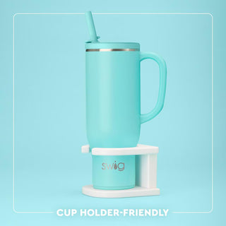 A mint green Swig travel mug with a handle, straw, and a white cup holder base, shown against a solid light blue background with a white border and text reading 'CUP HOLDER-FRIENDLY'.