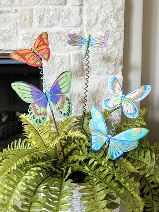 Colorful butterfly and dragonfly windsocks in a pot of ferns against a stone wall.