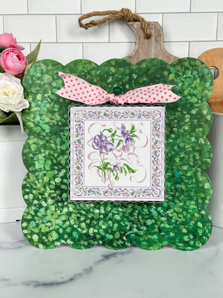 Decorative green scalloped frame with a pink bow, featuring a framed floral design on a white tiled wall background.