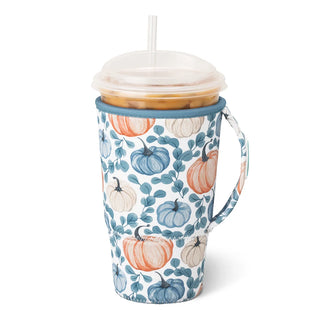 A handled insulated travel tumbler with a clear lid and straw, featuring a white fabric sleeve printed with blue and orange pumpkins and teal leaves. The tumbler is filled with an iced coffee and photographed on a clean white background.