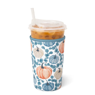 A clear plastic travel tumbler with a clear lid and straw, filled with an iced orange drink, wrapped in a white fabric sleeve printed with blue leaves and orange and beige pumpkins, on a white background.