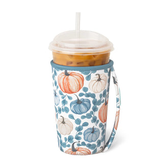 A handled insulated travel tumbler with a clear lid and straw, filled with an iced coffee. The tumbler is wrapped in a white fabric sleeve decorated with a pattern of blue, orange, and cream pumpkins and blue leaves.