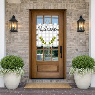 A decorative white scalloped 'Welcome' sign with a subtle plaid pattern and olive green leaf motif, displayed on a wooden front door. The door is set in a brick entryway flanked by two black lantern wall sconces and two large white planters with trimmed green bushes and white flowers.
