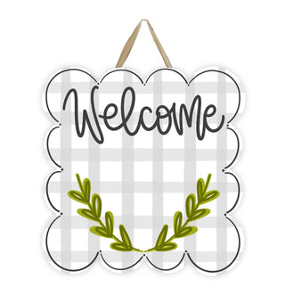 A scalloped white welcome sign with a gray buffalo plaid pattern, black script text reading 'Welcome,' and an olive green leafy wreath at the bottom. The sign has a hanging loop at the top and is shown on a plain white background.