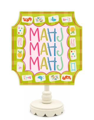 A decorative table lamp with a scalloped sign panel featuring the letters 'MAHJ' in pink, yellow, blue, and green on a pink background, surrounded by a green gingham border and small colorful tile illustrations. The lamp has a cream-colored pedestal base.