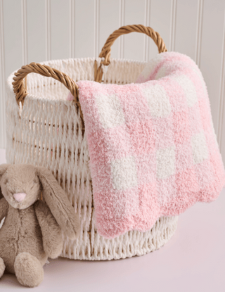 A pink and white checkered plush throw blanket draped over a woven cream basket with rope handles, styled with a beige stuffed bunny on a pale pink surface against a white paneled wall.