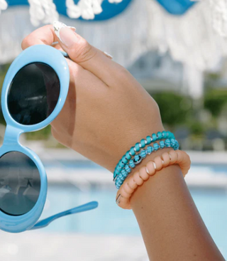 Hand holding blue sunglasses with a blurred pool and deck chair in the background