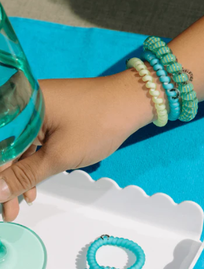 Hand holding a green bottle with turquoise bracelets on a blue surface
