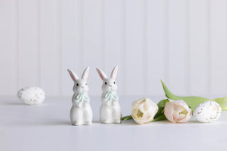 Two white ceramic bunny figures with green bows on a light gray background with flowers.