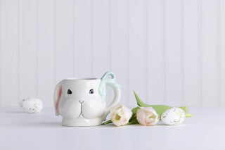 White ceramic mug shaped like a rabbit with pink ears, sitting on a light gray surface with white flowers and green leaves.