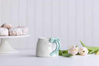 White mug with a turquoise handle on a light surface with flowers and pastries in the background