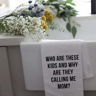 Towel with text 'WHO ARE THESE KIDS AND WHY ARE THEY CALLING ME MOM?' on a white surface with flowers.