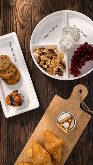 A styled flat-lay of children's feeding accessories on a dark wood table, including a white divided plate with printed text, a white rectangular tray with cookies and a small jar, a wooden cutting board, and pastries.