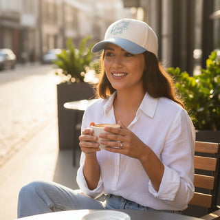 Woman sitting outdoors holding a coffee cup, wearing a cap and white shirt.