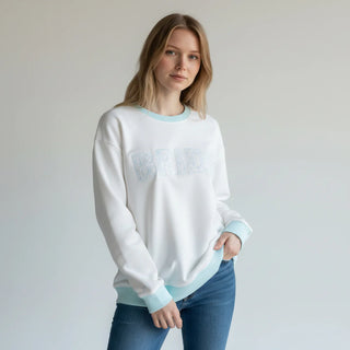 Woman wearing a white sweatshirt with light blue accents and jeans on a plain background