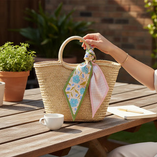 Person holding a straw bag with a floral scarf, next to a cup and notebook on a wooden table outdoors.