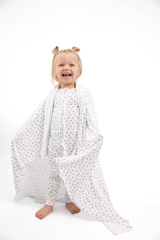 A smiling young child stands barefoot on a white studio background, wrapped in a white muslin blanket with a small gray pattern. The child has blonde hair styled in two buns and is wearing a light pink top.