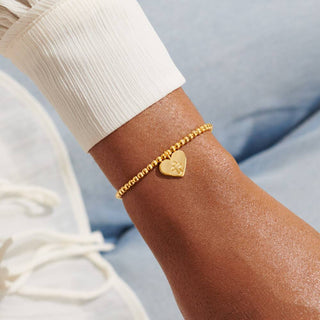Gold bracelet with a heart-shaped charm on a wrist against a light blue background