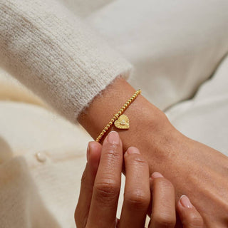 Gold bracelet with a heart charm on a wrist against a neutral background