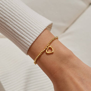 Gold bracelet with a heart charm on a wrist against a neutral background