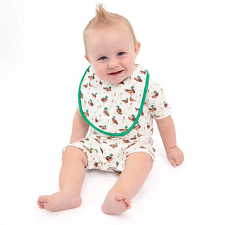 A baby sitting on a white studio background wearing a short-sleeve white romper with a brown duck print and a green-trimmed bib.