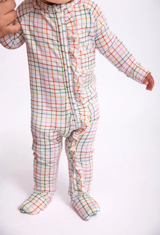 A baby wearing a long-sleeve footed sleeper with a pastel plaid pattern and ruffle trim down the front, standing against a plain white background.
