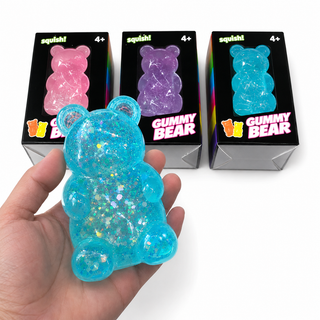 A hand holds a translucent blue glittery gummy bear in the foreground, with three boxed gummy bear products behind it on a white background. The boxes are black with colorful gummy bear images and the brand name 'squish!' visible.