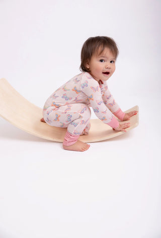 A baby wearing a light pink long-sleeve pajama set with a colorful butterfly print is crawling on a curved beige wooden board against a plain white studio background.