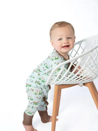 A baby wearing a light green bunny-print footless romper and brown booties stands behind a white wire chair on a wooden leg against a white studio background.