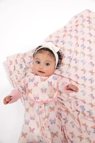 A baby wearing a white bow headband and a pink footed sleeper with a colorful butterfly print, lying on a matching butterfly-patterned blanket against a white background.