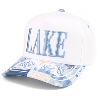 A white baseball cap featuring the word 'LAKE' embroidered in light blue on the front, with a blue and white nautical-themed patterned visor.