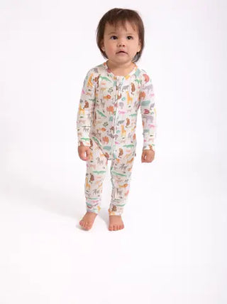 A toddler standing barefoot against a plain white studio background, wearing a long-sleeve footless one-piece pajama or romper with an allover colorful animal print on a light base.