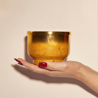 A hand with red manicured nails holds a small metallic gold container with a textured, hammered finish against a warm beige background.