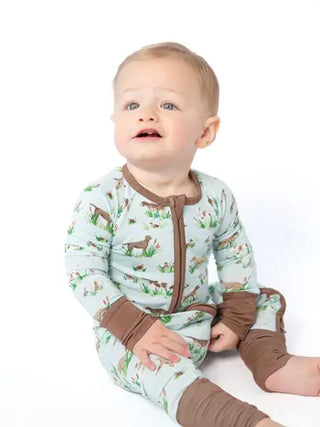 A baby sitting on a white background wearing a long-sleeve footless pajama romper with a brown zipper, brown cuffs and leg warmers, and a light green print of deer and grass.