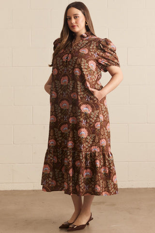 A woman modeling a brown midi dress with a colorful floral and paisley print, featuring puff sleeves and a tiered hem, standing against a beige brick wall.