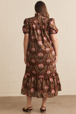 A woman viewed from behind wearing a brown midi dress with a colorful paisley and floral pattern, puff sleeves, and a tiered hem, standing against a light beige brick wall.