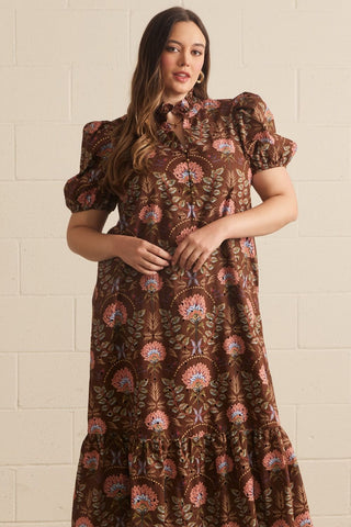 Woman wearing a brown floral dress against a beige tiled wall.